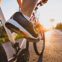 Nahaufnahme eines Radfahrers aus Höhe der Pedalen, auf einer Straße dem Sonnenuntergang entgegen.